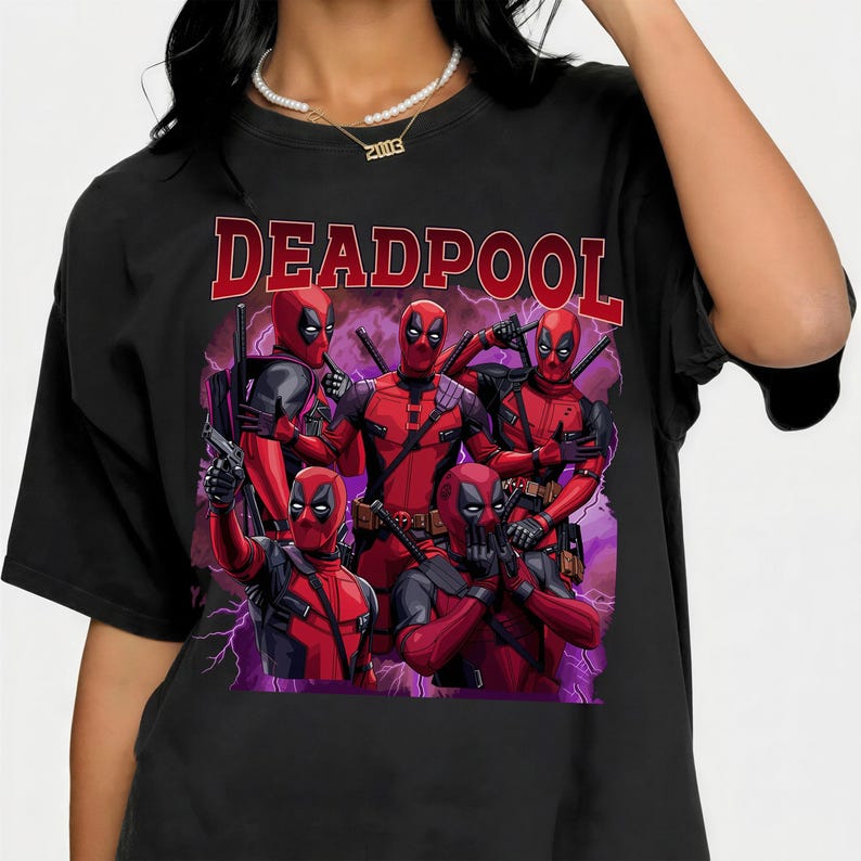 May include: Black t-shirt featuring a graphic of Deadpool characters in red and black costumes, with the word "DEADPOOL" in red block letters across the top. The design includes a purple lightning effect.