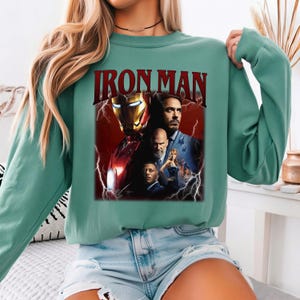 Pu&ograve; includere: Una felpa verde acqua con una grafica del poster del film Iron Man. Il poster include immagini del casco di Iron Man, Tony Stark e altri personaggi, con il titolo "IRON MAN" in lettere rosse.