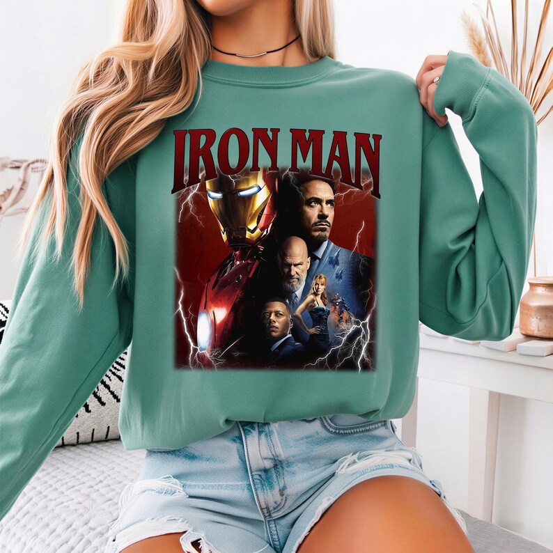 Pu&ograve; includere: Una felpa verde acqua con una grafica del poster del film Iron Man. Il poster include immagini del casco di Iron Man, Tony Stark e altri personaggi, con il titolo "IRON MAN" in lettere rosse.