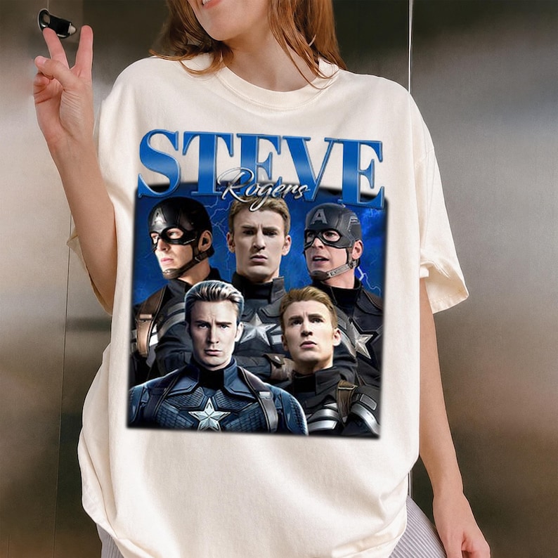 Op de afbeelding: Cr&egrave;mekleurig T-shirt met een afbeelding van Steve Rogers, met de tekst "STEVE Rogers" in blauw. De afbeelding bevat meerdere afbeeldingen van Captain America in verschillende poses, tegen een blauwe achtergrond.