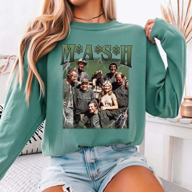 May include: A sage green crewneck sweatshirt featuring a graphic print of the M*A*S*H cast. The design includes the text M*A*S*H in a dark green font above a vintage-style photograph of the ensemble cast surrounded by white lightning bolt accents.