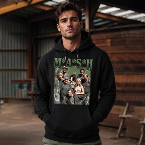 May include: A black hooded sweatshirt featuring a graphic print of the M*A*S*H cast. The design includes the green text M*A*S*H above a group photograph of the characters, surrounded by white lightning bolt illustrations on a dark green background.