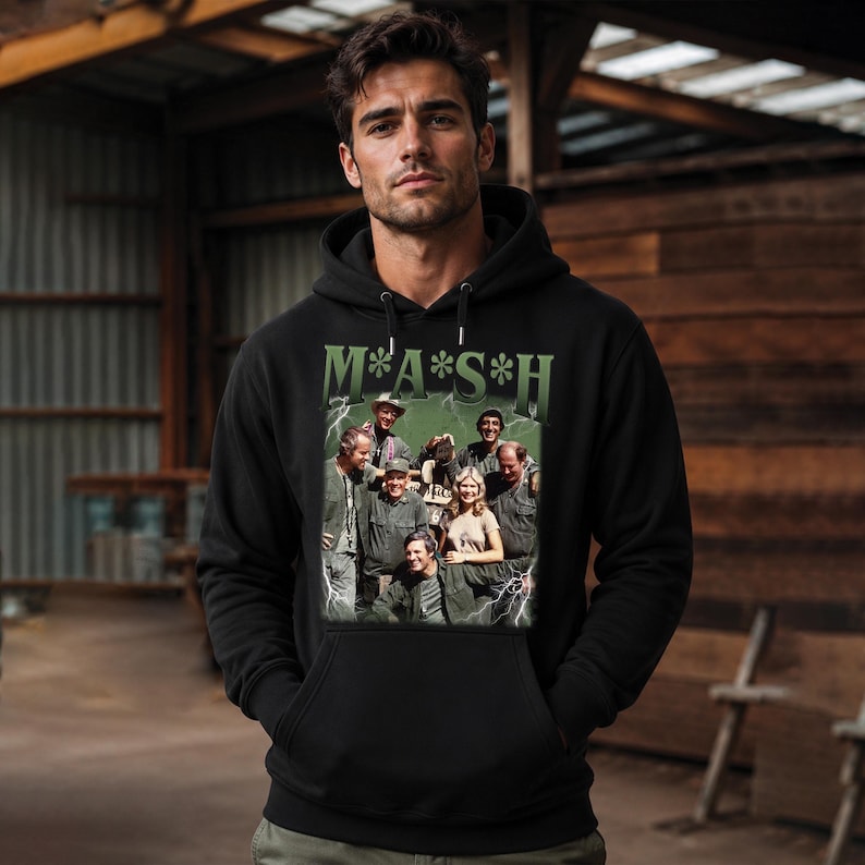 May include: A black hooded sweatshirt featuring a graphic print of the M*A*S*H cast. The design includes the green text M*A*S*H above a group photograph of the characters, surrounded by white lightning bolt illustrations on a dark green background.