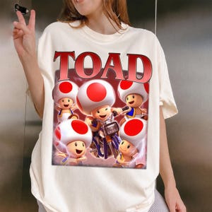 May include: Cream-colored t-shirt featuring a graphic of several cartoon Toads with red and white mushroom caps. One Toad is riding a motorcycle. The word "TOAD" is printed in red above the graphic.