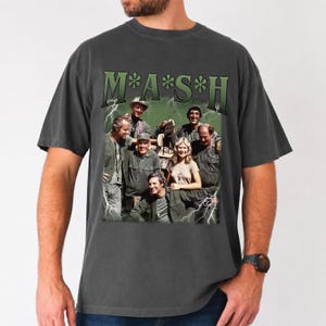 May include: A charcoal gray short-sleeve t-shirt featuring a graphic print of the M*A*S*H cast. The design includes the show title in green lettering at the top, a group photograph of the characters, and white lightning bolt accents on the sides.