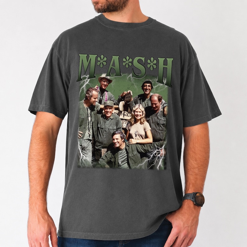 May include: A charcoal gray short-sleeve t-shirt featuring a graphic print of the M*A*S*H cast. The design includes the show title in green lettering at the top, a group photograph of the characters, and white lightning bolt accents on the sides.