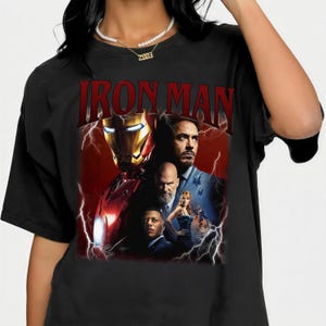 Pu&ograve; includere: T-shirt nera con una grafica di Iron Man e la scritta "IRON MAN" in rosso. Il design include immagini dell'armatura di Iron Man e dei personaggi del film, su uno sfondo di fulmini rossi e bianchi.