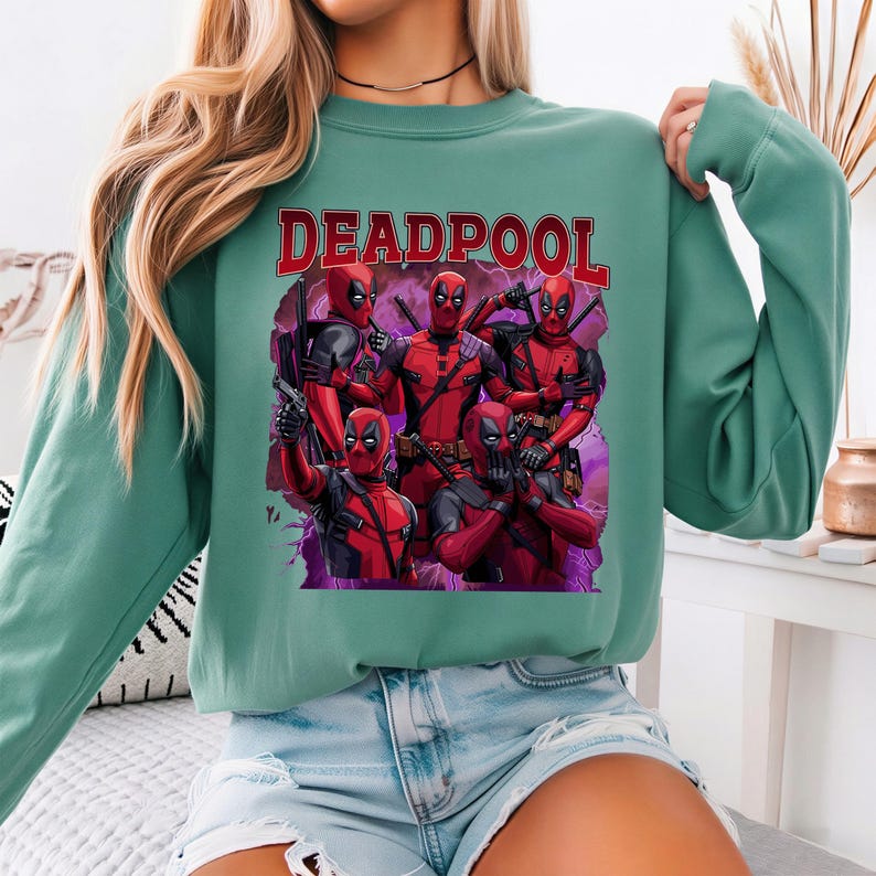 May include: A teal green sweatshirt featuring a graphic of Deadpool in various poses, with the word "DEADPOOL" in red block letters across the top. The graphic includes multiple figures of Deadpool in red and black costumes, set against a purple and black background.