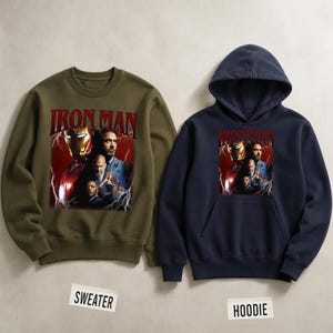 Pu&ograve; includere: Un maglione verde oliva e una felpa con cappuccio blu navy con una grafica di Iron Man. La grafica include il testo "IRON MAN" in rosso, insieme a immagini del personaggio e di altre figure. Gli indumenti sono presentati su uno sfondo neutro.