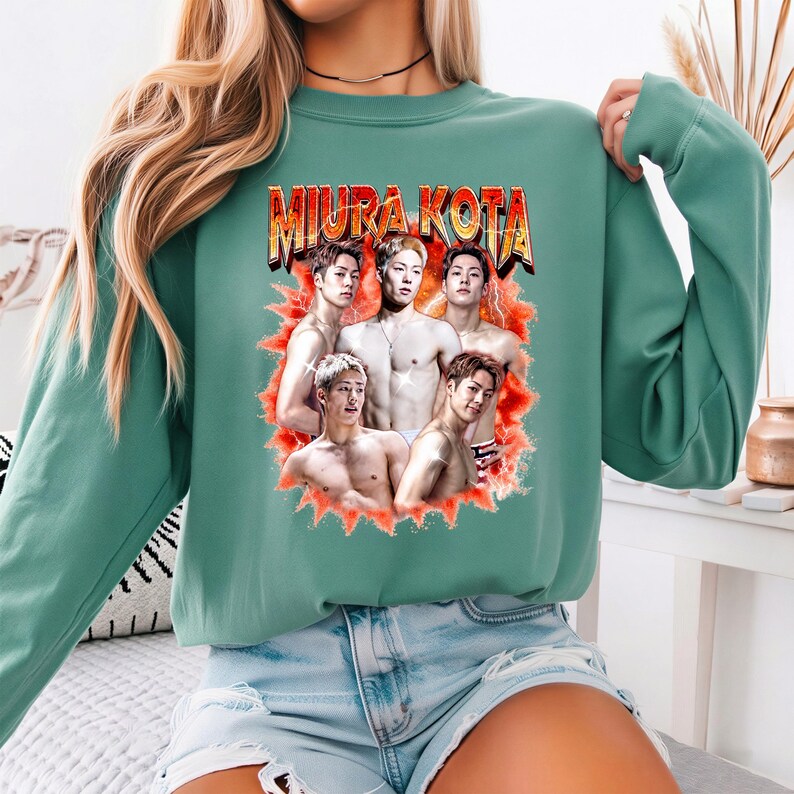 May include: A teal green sweatshirt featuring a graphic of five shirtless men with the text "MIURA KOTA" in a retro font. The graphic is surrounded by a red and orange burst design. The sweatshirt is a casual style.