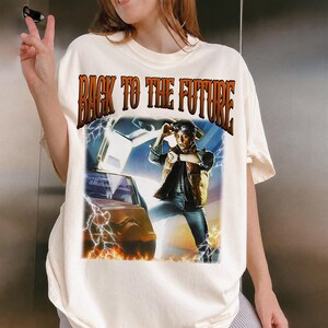 May include: Cream-colored t-shirt featuring a graphic from the movie "Back to the Future." The design includes the text "BACK TO THE FUTURE" in orange lettering, along with an image of a character and a car with lightning effects.
