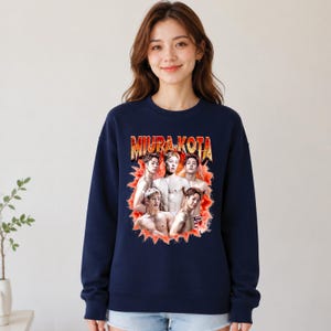 May include: Navy blue sweatshirt featuring a graphic of a group of men with the text "MIURA KOTA" in a retro style. The graphic is in shades of red, orange, and yellow. The sweatshirt is a crew neck style.