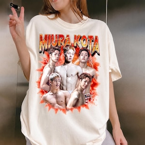 May include: Cream-colored t-shirt featuring the text "MIURA KOTA" in a gold and red design. The shirt displays a graphic of five shirtless men with a red lightning bolt background.