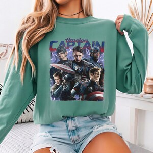 May include: A teal green sweatshirt featuring a graphic of Captain America with the text "America Captain". The graphic shows multiple images of Captain America in different poses. The sweatshirt has long sleeves and a crew neck.