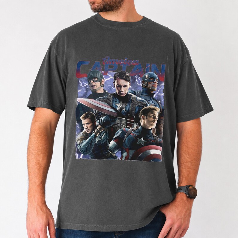 May include: Dark gray t-shirt featuring a graphic of Captain America and other characters. The design includes the words "American Captain" in a stylized font, with lightning bolts in the background. The shirt is a crew neck.