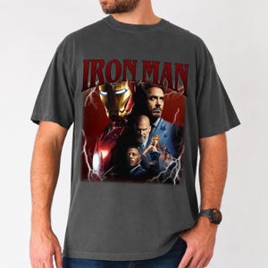 Pu&ograve; includere: T-shirt grigio scuro con la scritta rossa "IRON MAN" sopra una grafica dell'armatura di Iron Man e diversi personaggi. La grafica ha uno sfondo rosso con fulmini.