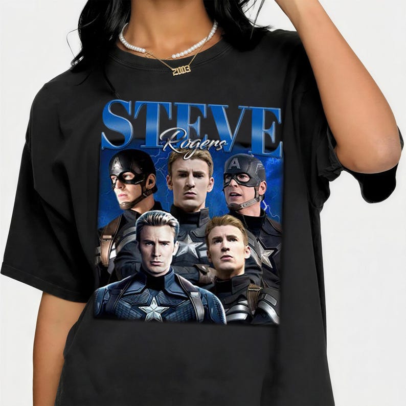 Op de afbeelding: Zwart T-shirt met een afbeelding van Steve Rogers uit de Captain America-films. Het ontwerp bevat de naam "Steve Rogers" in blauw, met meerdere afbeeldingen van het personage in verschillende kostuums.