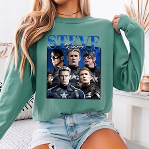 Op de afbeelding: Een teal-groene sweatshirt met een afbeelding van Steve Rogers uit de Captain America-films. De afbeelding bevat meerdere afbeeldingen van het personage en de tekst "STEVE Rogers" in een blauw, gestileerd lettertype. De sweatshirt heeft een ronde hals en lange mouwen.