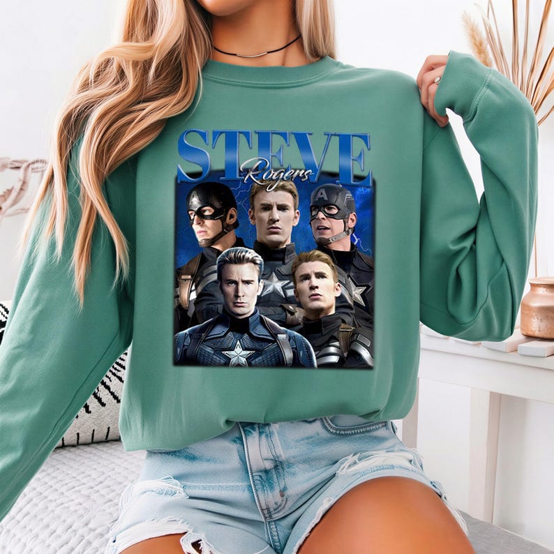Op de afbeelding: Een teal-groene sweatshirt met een afbeelding van Steve Rogers uit de Captain America-films. De afbeelding bevat meerdere afbeeldingen van het personage en de tekst "STEVE Rogers" in een blauw, gestileerd lettertype. De sweatshirt heeft een ronde hals en lange mouwen.