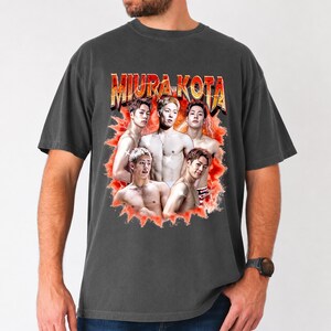 May include: Dark gray t-shirt featuring a graphic of five men with the text "MIURA KOTA" in a retro style. The graphic is set against a red and orange burst design. The shirt is a casual style.