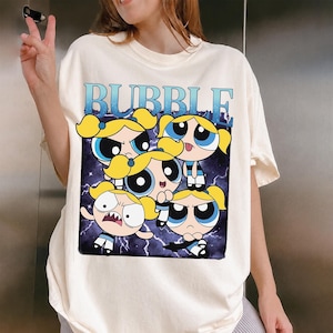 May include: Cream-colored t-shirt featuring a graphic of the Powerpuff Girls with the word "BUBBLE" in blue. The cartoon characters have yellow hair and large eyes, set against a dark blue background with lightning bolts.