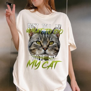 May include: Cream-colored t-shirt featuring a close-up of a cat's face with green eyes. The shirt has the text "DON'T TELL ME WHAT TO DO, YOU'RE NOT MY CAT" in a mix of black and green fonts.