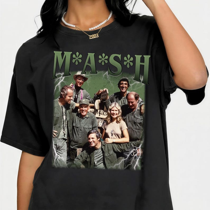May include: A black short-sleeved t-shirt featuring a large graphic print of the M*A*S*H cast. The design includes the green text M*A*S*H at the top, a group photograph of the characters, and white lightning bolt accents surrounding the central image.