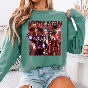 Puede incluir: Una sudadera verde azulado con un gr&aacute;fico de Iron Man en varias poses. La palabra "IRON MAN" est&aacute; impresa en letras rojas. El gr&aacute;fico incluye im&aacute;genes de la cara y la armadura de Iron Man.