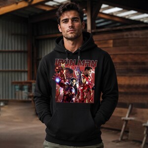 Puede incluir: Sudadera con capucha negra con un gr&aacute;fico de Iron Man en varias poses y el texto "IRON MAN". La sudadera tiene un bolsillo delantero y una capucha con cord&oacute;n. El fondo es una estructura de madera.