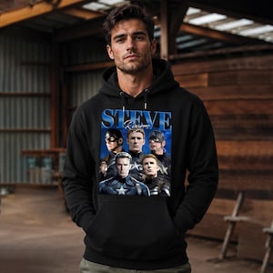 Op de afbeelding: Zwarte hoodie met een afbeelding van Steve Rogers in verschillende Captain America-poses. De tekst "STEVE Rogers" is blauw. De hoodie heeft een voorzak en een capuchon met trekkoord.
