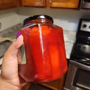 Pode incluir: Um frasco de vidro transparente cheio de pimentos vermelhos assados, com uma tampa preta de rosca. A superfície do vidro apresenta texto impresso, incluindo 'VCX50 01-024-PRO' e 'BEST IF USED BY SEP 25 2025', além de um rótulo parcial na lateral.