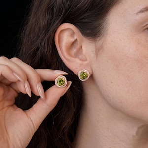 May include: A pair of rose gold stud earrings featuring a round, faceted lime green peridot gemstone at the center, surrounded by a delicate halo of small, clear white pavé stones arranged in a swirling, circular metal frame design.