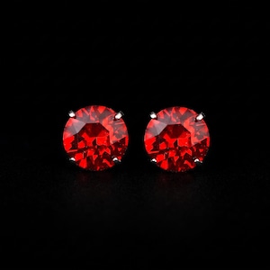 May include: A pair of round stud earrings featuring vibrant red faceted stones. Each circular gemstone is secured by four small silver-toned metal prongs, creating a classic solitaire design that catches the light against a dark background.