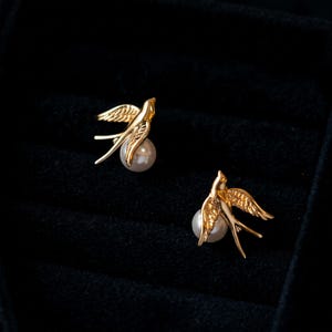 May include: A pair of gold-toned swallow bird earrings, each featuring a polished white spherical pearl held within the bird's wings. The swallows are depicted in flight with detailed wing feathers and long, pointed tail feathers, showcasing a metallic finish.