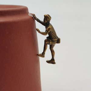 Puede incluir: Una pequeña figura de color bronce de un escalador sujeta al lateral de una taza de cerámica de color terracota. El escalador aparece en una pose dinámica con una mano extendida hacia arriba y los pies apoyados sobre la superficie curva.