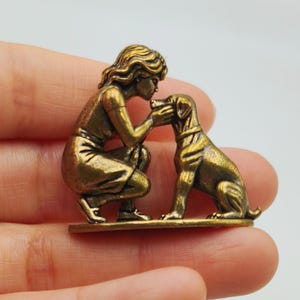 May include: A small, antique-style brass-toned metal figurine depicting a person kneeling and interacting with a dog. The figure shows a person with wavy hair wearing a dress, reaching towards the dog's head, while the dog sits upright.