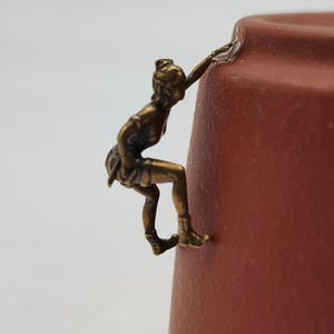 May include: A small bronze-colored metal figurine of a climber attached to the side of a matte terracotta-colored ceramic vessel. The climber is depicted in a dynamic pose with one hand reaching upward and feet braced against the curved surface.