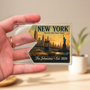 May include: A rectangular clear plastic luggage tag insert featuring a New York City skyline illustration with the Statue of Liberty, yellow taxis, and the text NEW YORK The City That Never Sleeps, The Johnsons, and Est. 2026 in dark blue lettering.