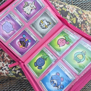 May include: A bright pink zipped binder containing eight trading cards arranged in clear plastic sleeves. Each card features a colourful cartoon character illustration, the text Basic Energy, and a small signature in the bottom corner.