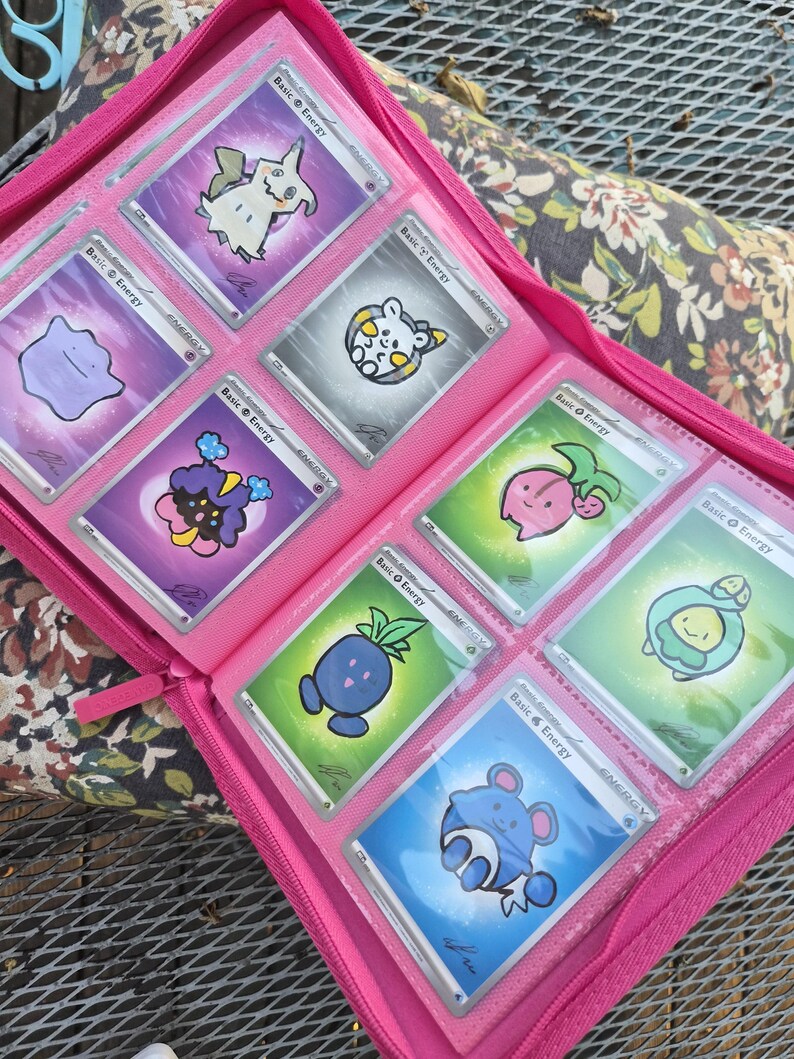 May include: A bright pink zipped binder containing eight trading cards arranged in clear plastic sleeves. Each card features a colourful cartoon character illustration, the text Basic Energy, and a small signature in the bottom corner.