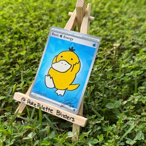 May include: A trading card featuring a yellow cartoon duck character on a blue background, labelled Basic Energy, displayed on a small wooden easel. The card includes the text 0054/01 and a signature, while the easel is marked @Poke Palette Binders.