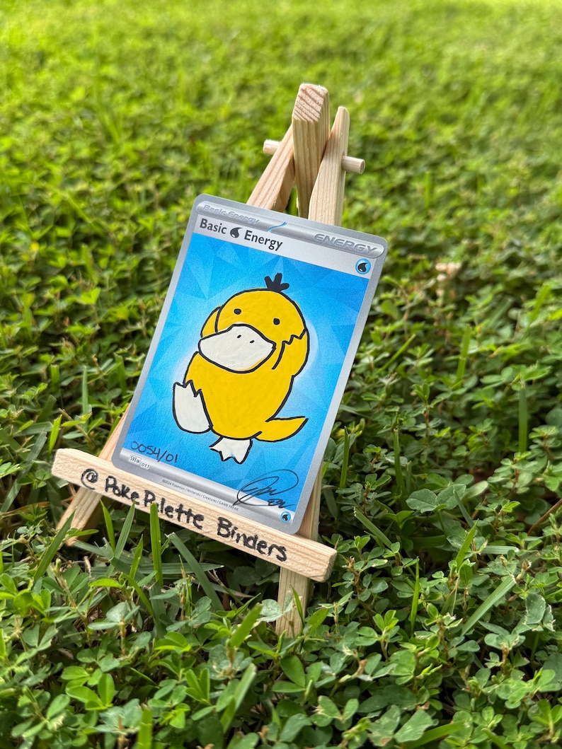 May include: A trading card featuring a yellow cartoon duck character on a blue background, labelled Basic Energy, displayed on a small wooden easel. The card includes the text 0054/01 and a signature, while the easel is marked @Poke Palette Binders.