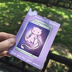 May include: A light purple card holder featuring a small white rabbit icon at the top, the text Pok&eacute;Palette Binders, and the text Thank you for your order! at the bottom. A psychic energy trading card with a pink Mew illustration is inserted inside.