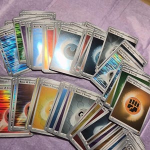May include: A collection of assorted Pok&eacute;mon trading cards featuring various Basic Energy types. The cards display colourful symbols including water droplets, lightning bolts, fire flames, leaves, and fighting fists, with some cards showing a holographic finish.