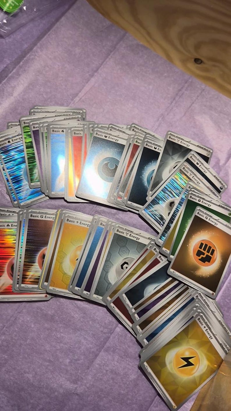 May include: A collection of assorted Pok&eacute;mon trading cards featuring various Basic Energy types. The cards display colourful symbols including water droplets, lightning bolts, fire flames, leaves, and fighting fists, with some cards showing a holographic finish.