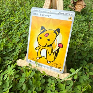 May include: A rectangular trading card featuring a yellow background with the text Basic Energy and a lightning bolt symbol. A hand-drawn illustration of a yellow creature with black stripes and a red tail tip is centred on the card.