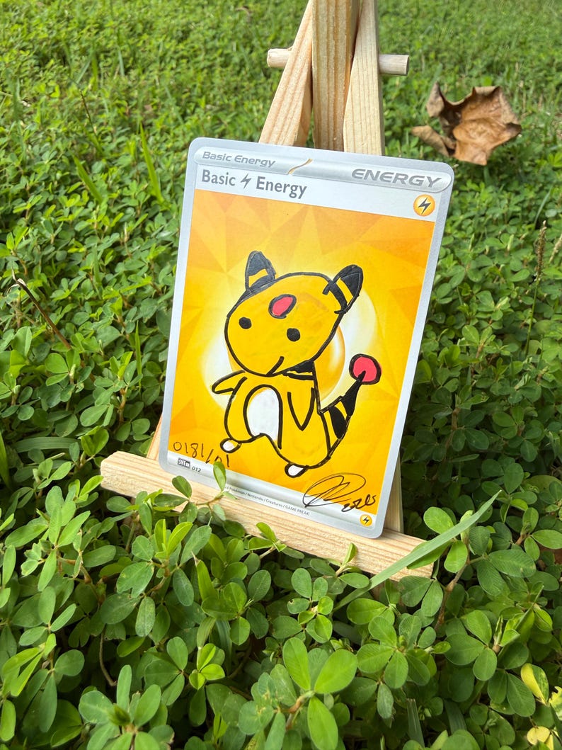 May include: A rectangular trading card featuring a yellow background with the text Basic Energy and a lightning bolt symbol. A hand-drawn illustration of a yellow creature with black stripes and a red tail tip is centred on the card.