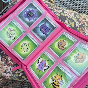 May include: A bright pink zipped trading card binder containing eight individual trading cards. Each card features a unique cartoon creature illustration, including a purple ghost, a yellow bee, and various green-themed characters, all labelled Basic Energy.