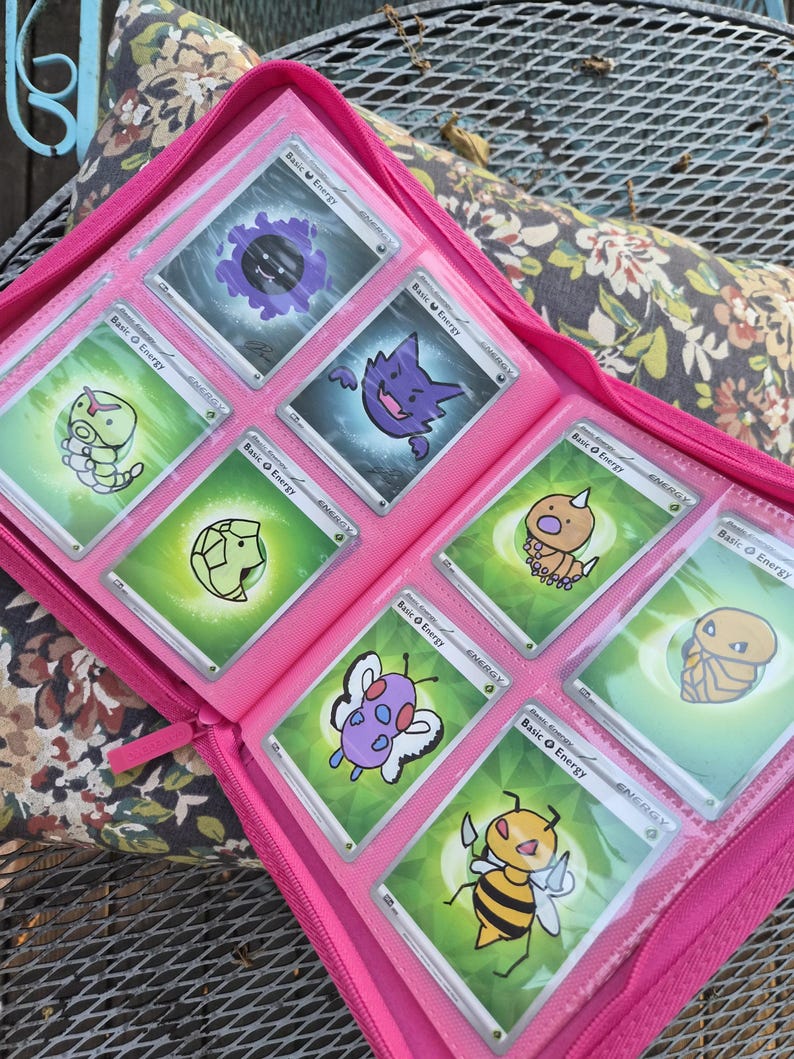 May include: A bright pink zipped trading card binder containing eight individual trading cards. Each card features a unique cartoon creature illustration, including a purple ghost, a yellow bee, and various green-themed characters, all labelled Basic Energy.