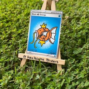 May include: A rectangular trading card featuring a blue background and a central orange fish illustration with yellow fins and whiskers. The text reads Basic Energy, Basic Water Energy, 2129/01, and a signature. The card rests on a small wooden easel.